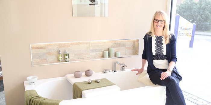 In einem hellen Badezimmer posiert eine Frau in eleganter Kleidung, während sie auf der Badewanne sitzt und freundlich in die Kamera lächelt.