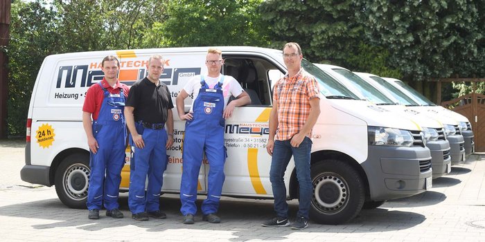 Vier Handwerker in blauen Overalls stehen lächelnd vor einem Firmenfahrzeug, das für Heizungs- und Sanitärdienstleistungen wirbt.